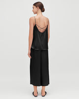 Woman standing, wearing a top with crossed back straps and loose pants, viewed from the back.