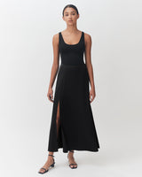 Woman in sleeveless top and long skirt standing against a light background