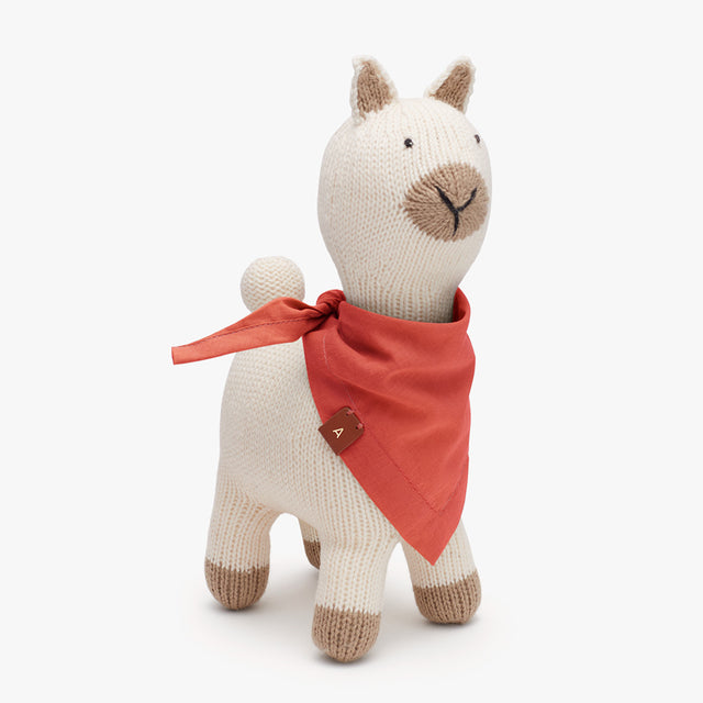 Stuffed toy alpaca with a bandana around its neck standing against a solid background.