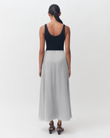 Woman standing facing away, wearing a sleeveless top and long skirt, with her hair tied back.