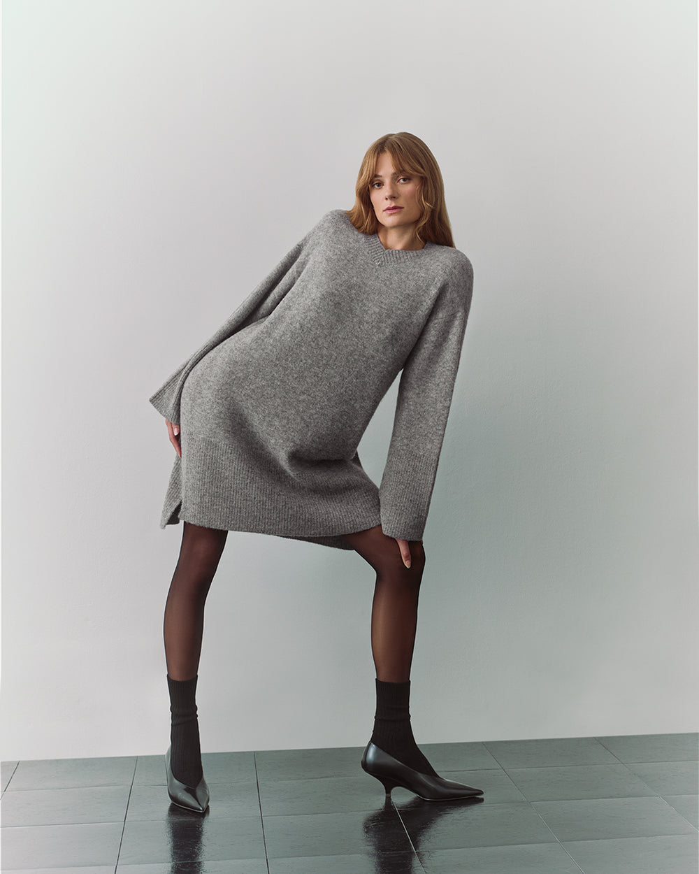 Person in oversized sweater dress and heels poses on tiled floor.