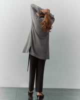 Person in long sweater and pants stands with arms raised, facing away.