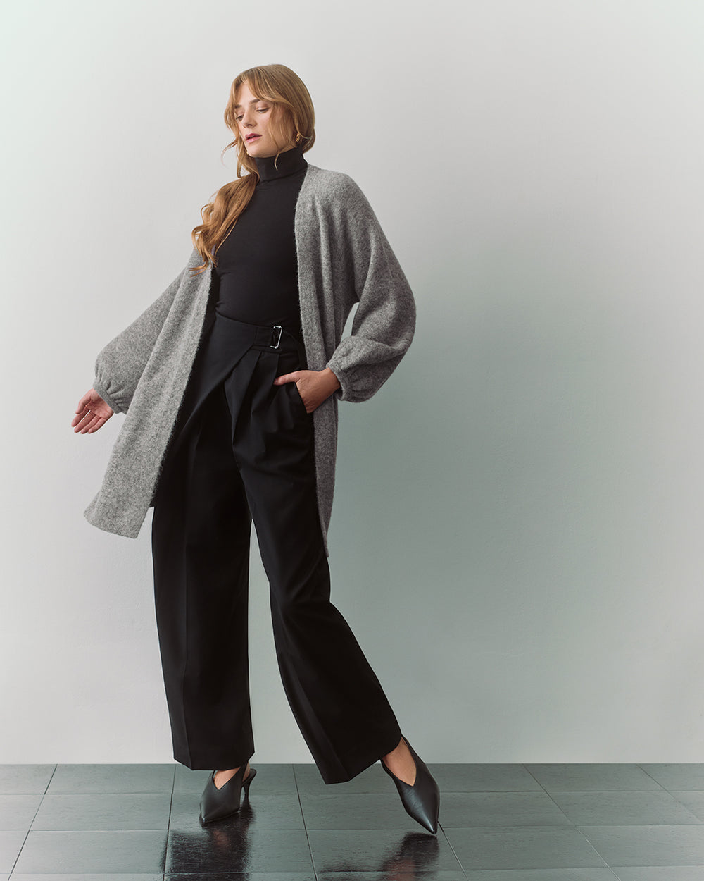 Person in long cardigan over turtleneck and wide-leg pants standing on tiled floor.