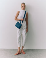 Person in sleeveless top and pants holding a handbag, wearing sandals standing against a wall.