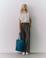 Person standing, wearing a top and trousers, holding a tote bag.