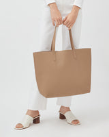 Person standing holding a large handbag, wearing heeled sandals and white pants.