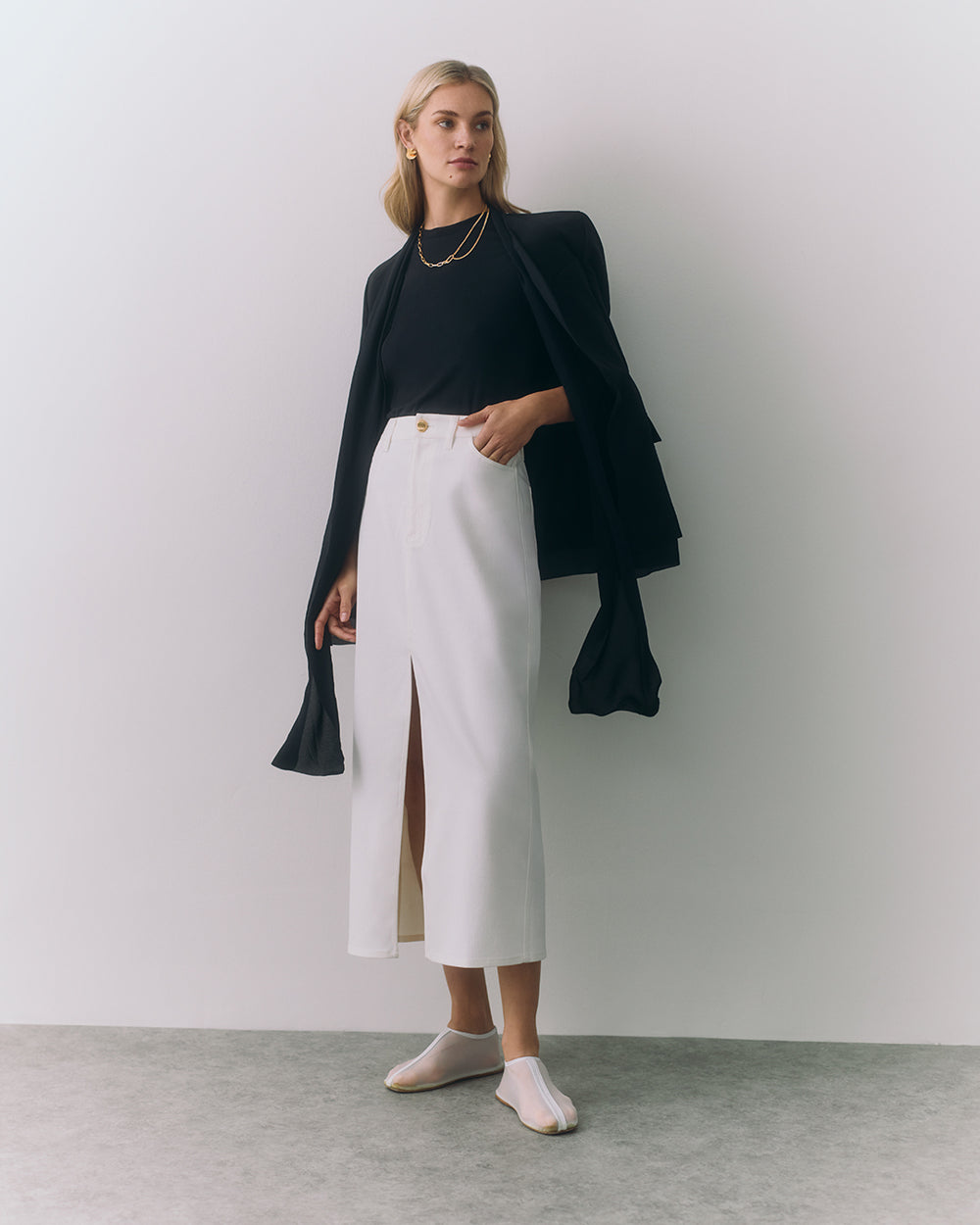 Woman in a long skirt, top, and jacket standing indoors with her hands in pockets.