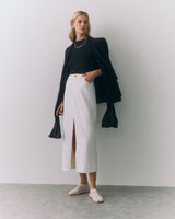 Woman in a long skirt, top, and jacket standing indoors with her hands in pockets.