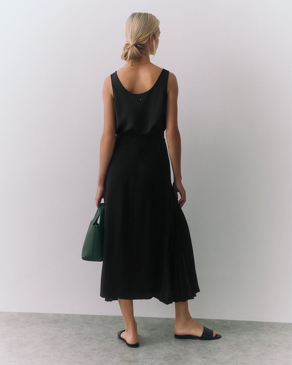 Person in a sleeveless top, long skirt, holding a handbag, wearing sandals, standing indoors.
