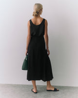 Person in a sleeveless top, long skirt, holding a handbag, wearing sandals, standing indoors.