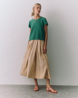 Woman in a short-sleeved top and long beige skirt wearing sandals standing against a wall.