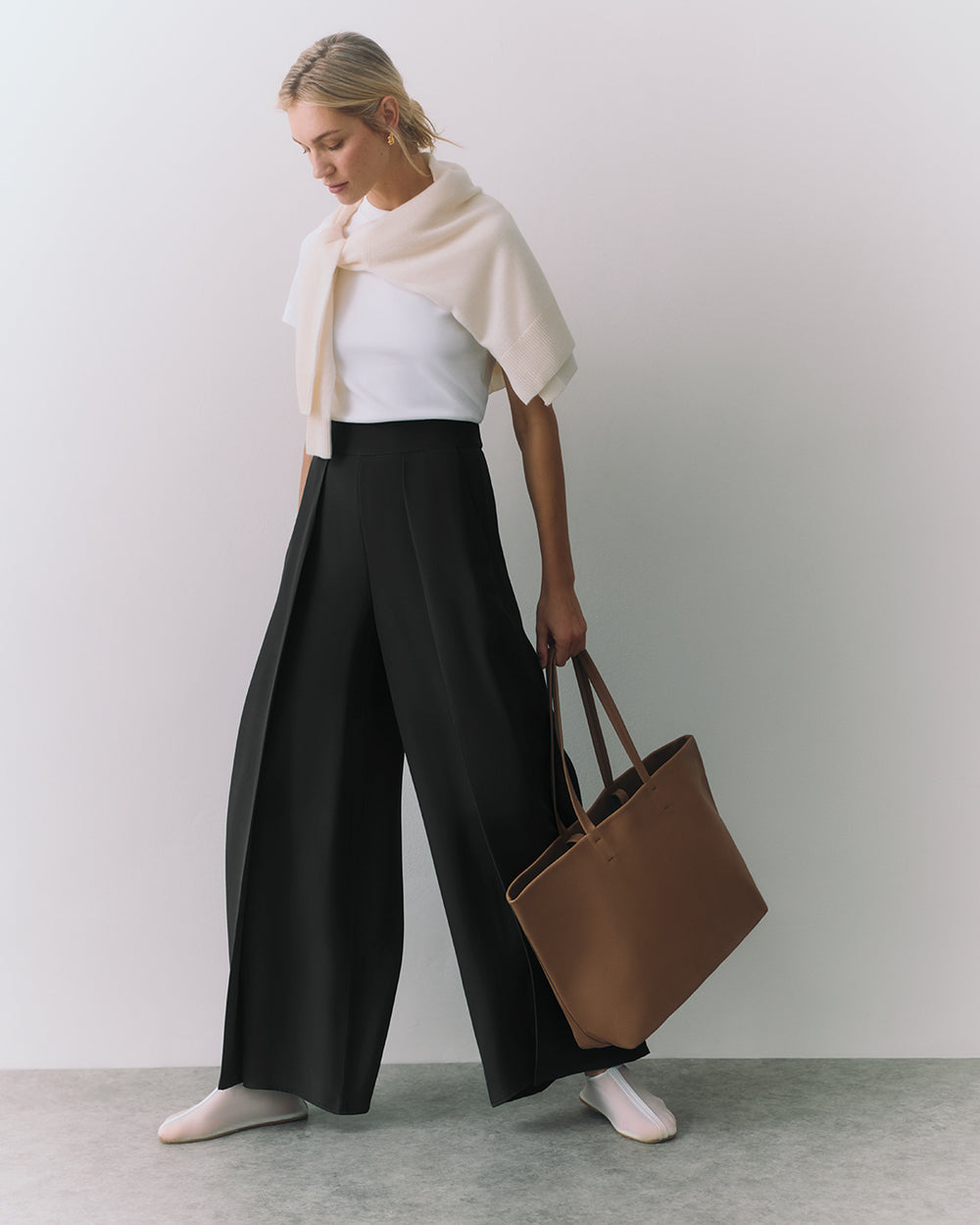 Person in wide trousers with a sweater over shoulders holding a large tote bag