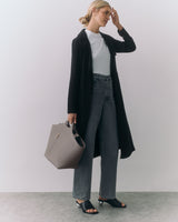 Person in long coat and jeans holding a large handbag standing indoors.