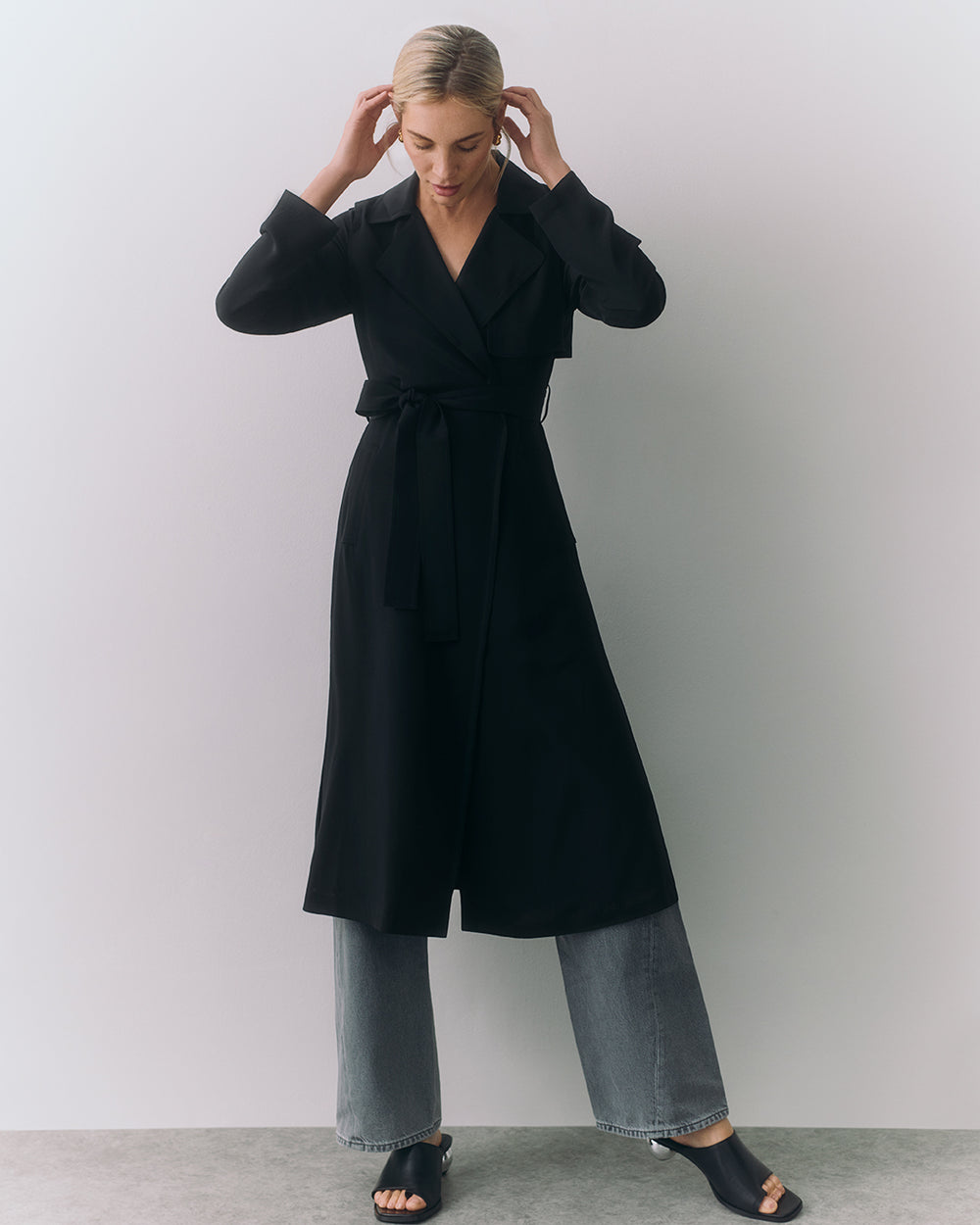 Person wearing long coat over wide-leg pants, standing with hands touching hair.