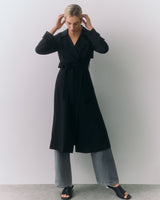 Person wearing long coat over wide-leg pants, standing with hands touching hair.