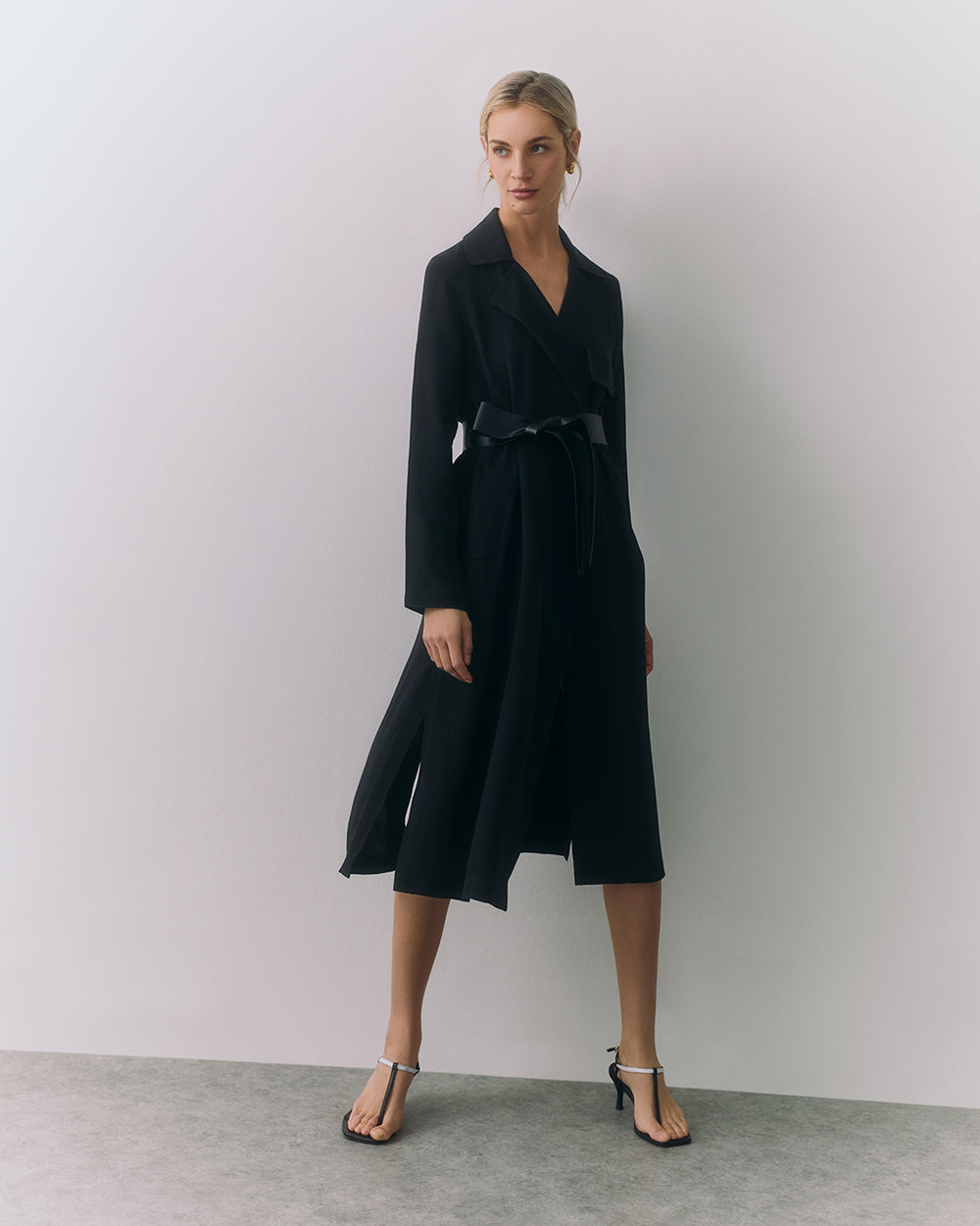 Person wearing a long coat with a belt, standing in a plain indoor setting.