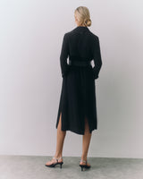 Person standing, facing a wall, wearing a long coat and heels.