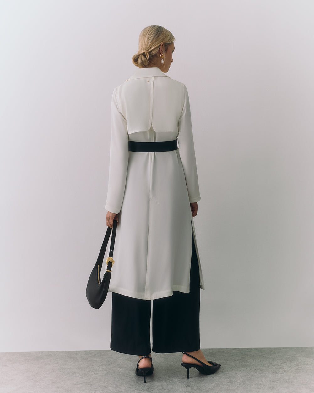 Woman in long coat and wide pants holds a handbag standing against plain background
