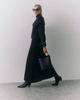 Person in long skirt, boots, holding a handbag, wearing sunglasses, walking indoors.