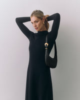 Person in long-sleeve dress holding a small shoulder bag against plain background.