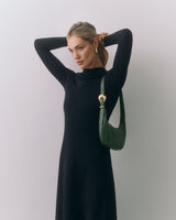 Person in long dress with a shoulder bag adjusts hair, standing against plain background.