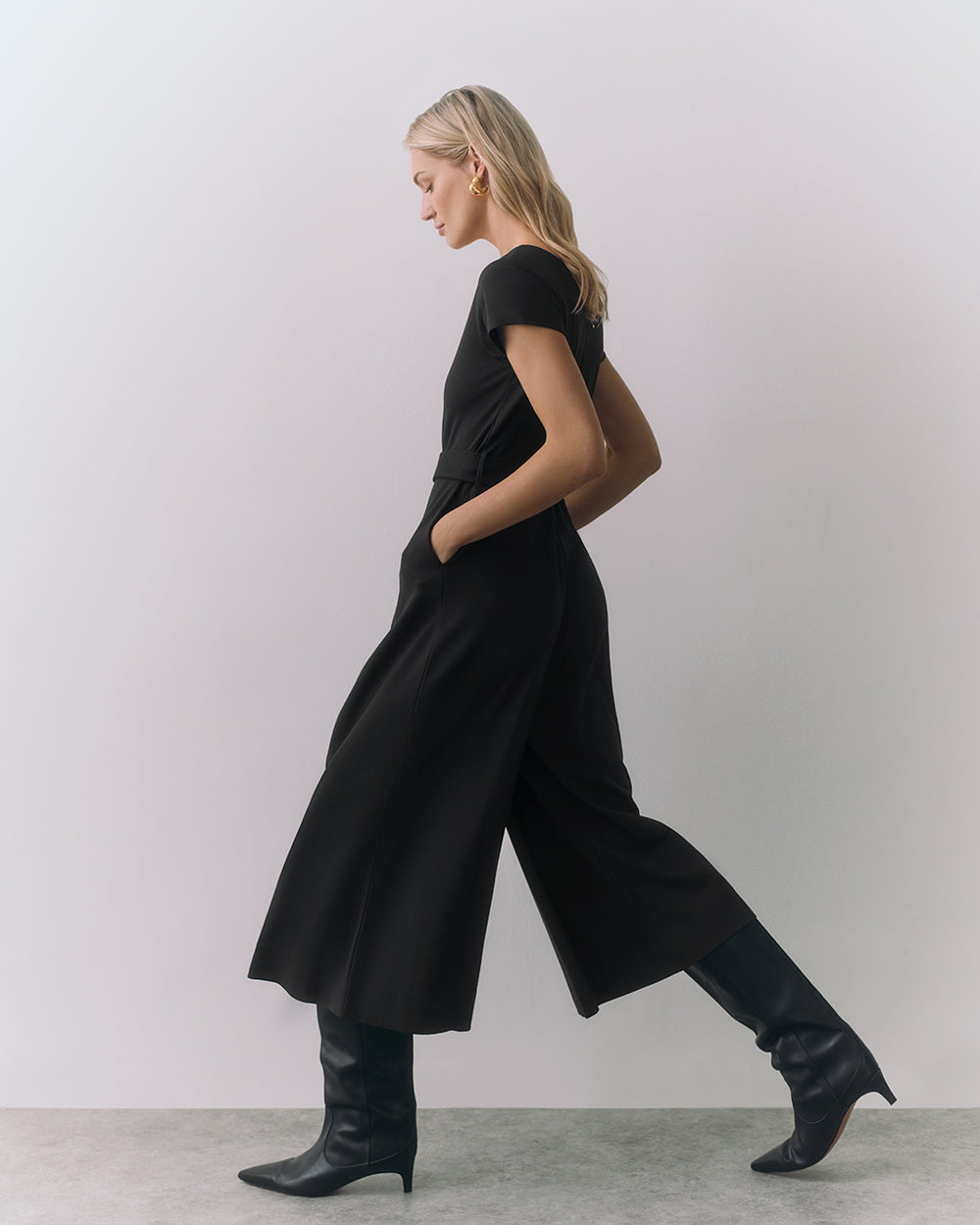 Ponte Pleated Jumpsuit Model walking in a dress and boots, hands in pockets, side view.
