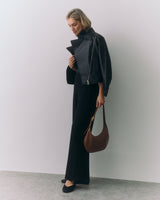Person in a jacket holding a handbag stands against a plain wall
