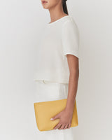 Woman holding a clutch, wearing a short-sleeved shirt and pants, standing against a plain background