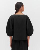 Woman seen from behind, wearing a top with voluminous sleeves and her hair in a bun.