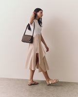 Woman in a skirt and sandals standing against a plain wall carrying a shoulder bag