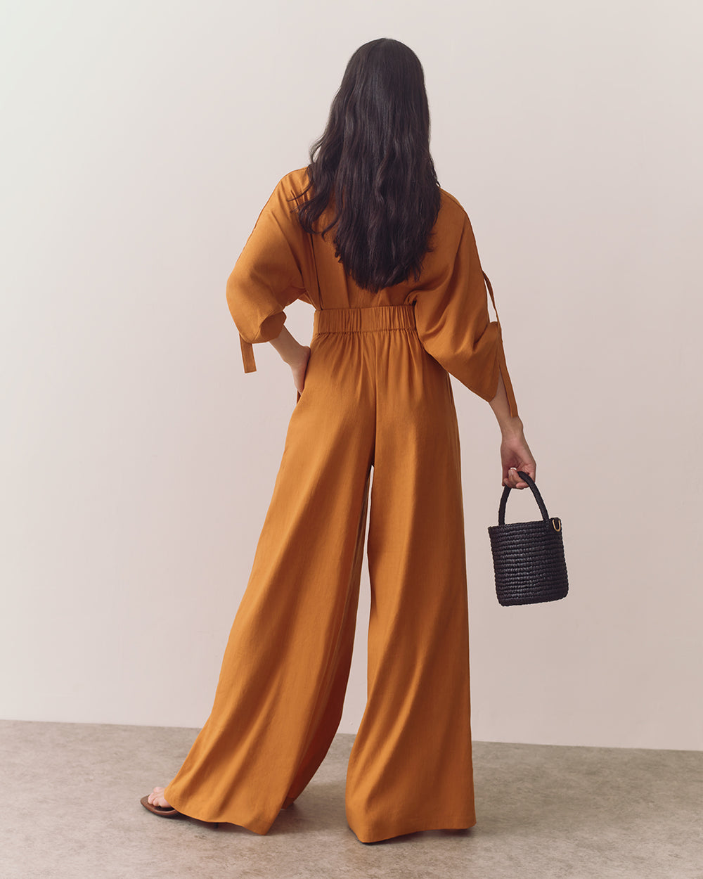 Person in wide-legged outfit holding small handbag in one hand, standing against plain backdrop.