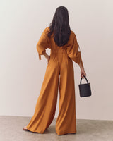 Person in wide-legged outfit holding small handbag in one hand, standing against plain backdrop.