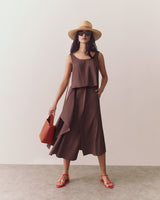 Person in a sleeveless top and flowy skirt holding a small handbag, standing indoors