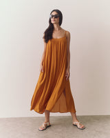 Person wearing a flowing dress, sandals, and sunglasses against a plain backdrop.