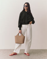 Person wearing sunglasses, jacket, pants, sandals, holding a handbag, stands indoors.