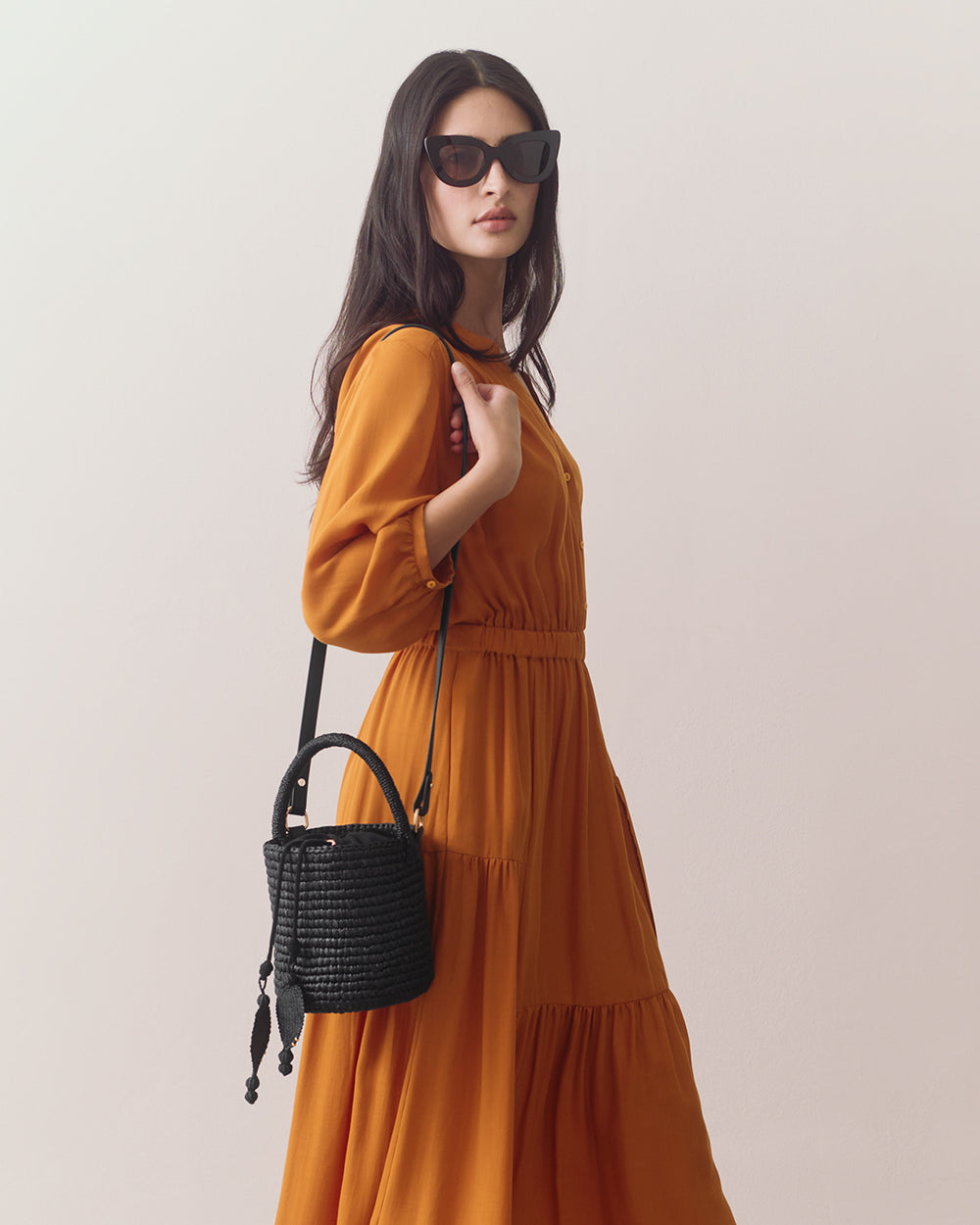 Person wearing sunglasses holds a woven bucket bag, dressed in a long, flowing dress.