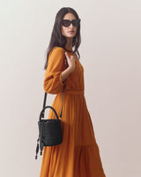Person wearing sunglasses holds a woven bucket bag, dressed in a long, flowing dress.