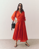 Woman in a long dress with sunglasses, holding a woven bag, standing indoors.