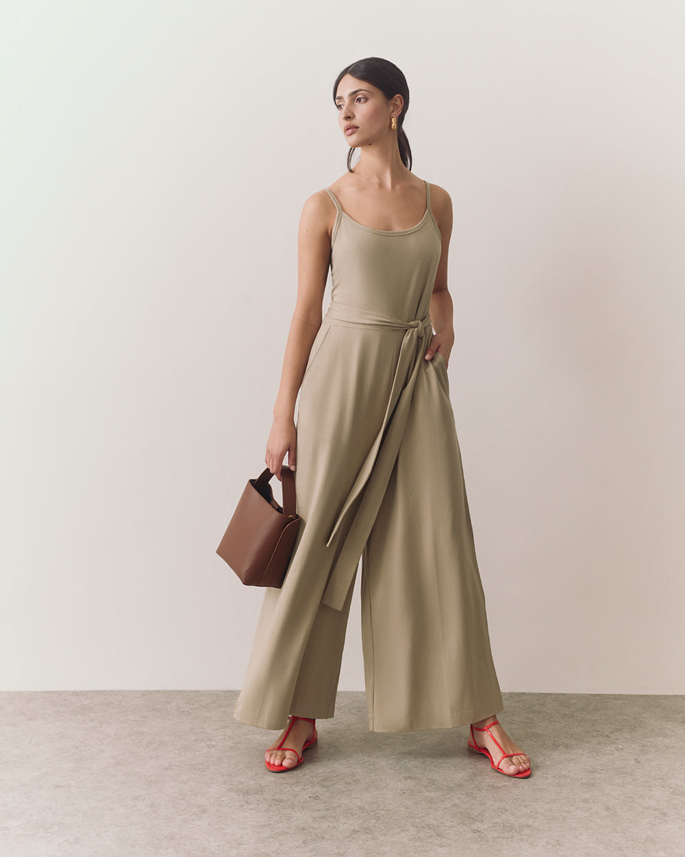 Person wearing jumpsuit with belt holds handbag and stands on a plain background.