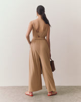 Person wearing a sleeveless jumpsuit, holding a handbag, standing on a plain background.