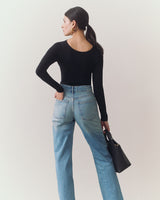 Person in jeans and long sleeve top holding handbag, facing away.