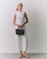 Person standing, holding a small bag, wearing a sleeveless top and pants, with sandals.