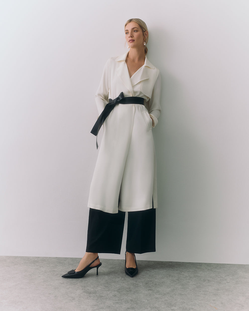 Woman in long coat with black belt and pants standing against a plain wall.