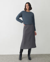 Woman in long skirt, boots, and long-sleeved top stands on carpeted floor.