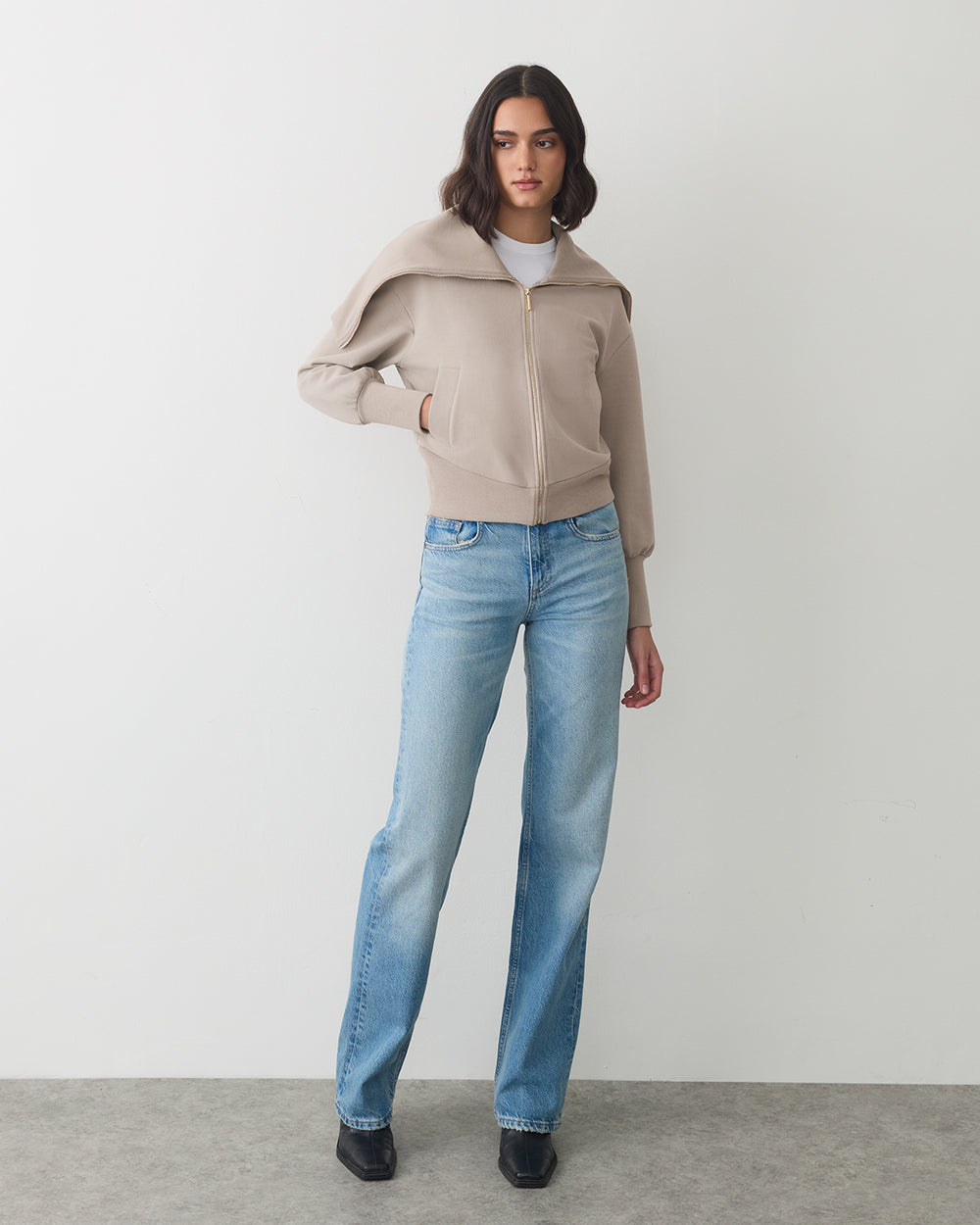 Person wearing a zip-up jacket, jeans, and shoes, standing against a plain backdrop.