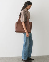Person standing with a shoulder bag, wearing a scarf, shirt, jeans, and boots.