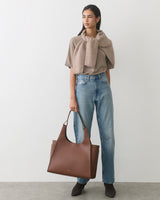 Person wearing jeans and top, carrying a large tote bag, stands against plain backdrop.
