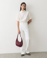 Person in white outfit holding a shoulder bag, standing against a plain wall.