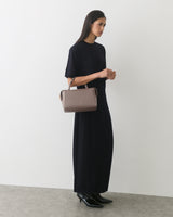 Person wearing long dress holding handbag, standing against plain wall.