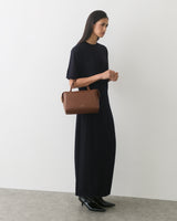Person in long dress holding a handbag, standing against a plain backdrop.
